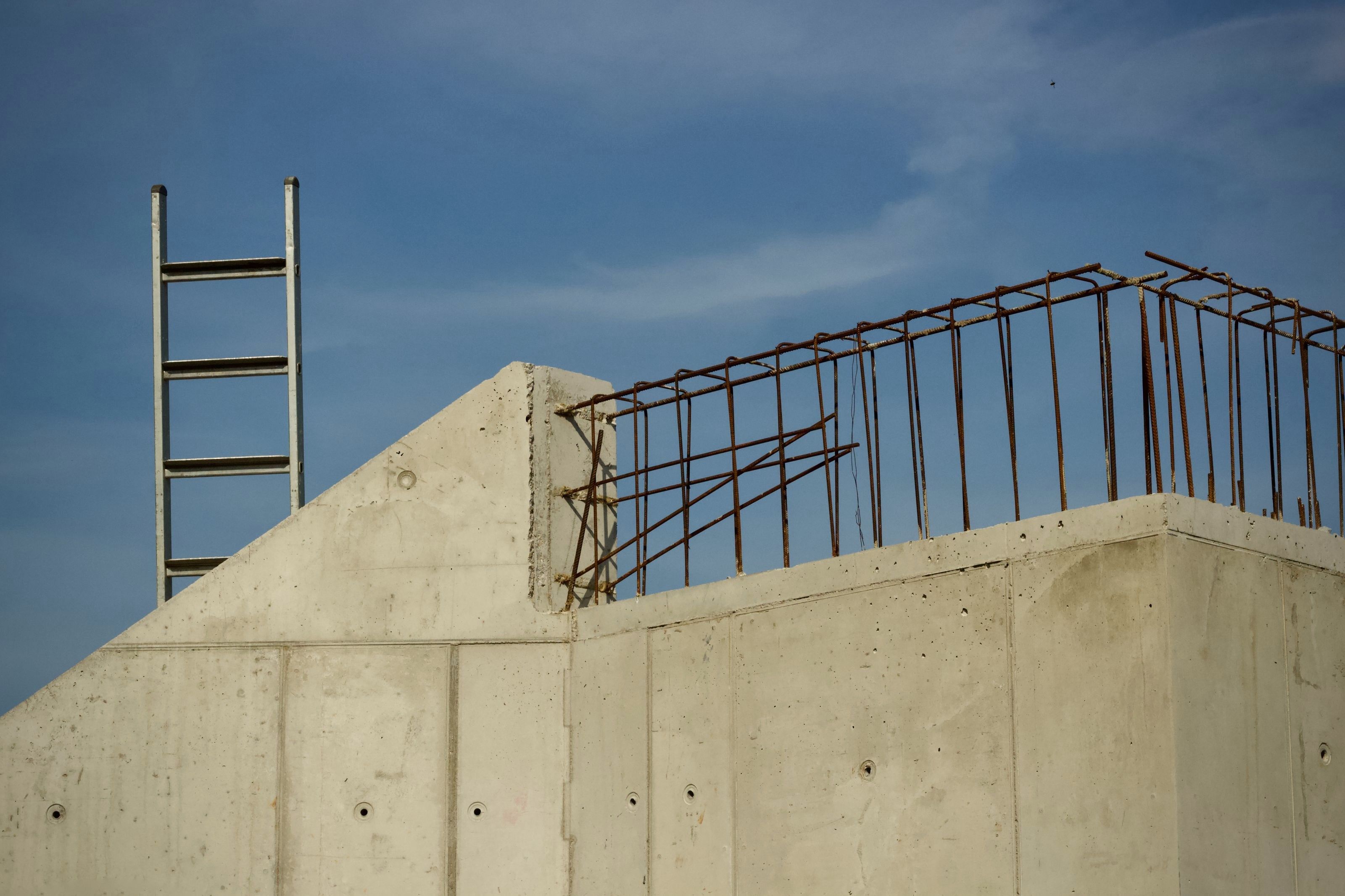 Gjuten betongvägg med utstickande armering och en stege mot blå himmel